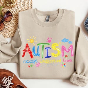 May include: Beige crewneck sweatshirt with the word "Autism" in colorful embroidered letters. The design includes a sun, clouds, handprints, and the words "accept, understand, love". The sweatshirt is paired with jeans and brown shoes.