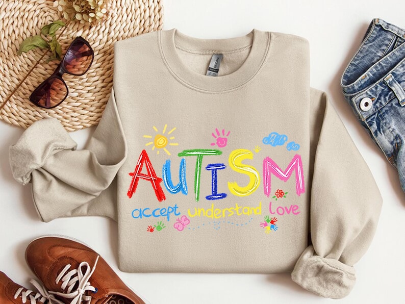 May include: Beige crewneck sweatshirt with the word "Autism" in colorful embroidered letters. The design includes a sun, clouds, handprints, and the words "accept, understand, love". The sweatshirt is paired with jeans and brown shoes.