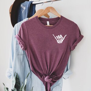 Op de afbeelding: Een maroon t-shirt met een witte skelethand die een shaka-teken maakt.
