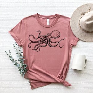 May include: Dusty rose t-shirt featuring a detailed black octopus illustration. The shirt is styled with a beige hat and a white mug. A sprig of greenery adds a decorative touch. The t-shirt is knotted at the bottom.
