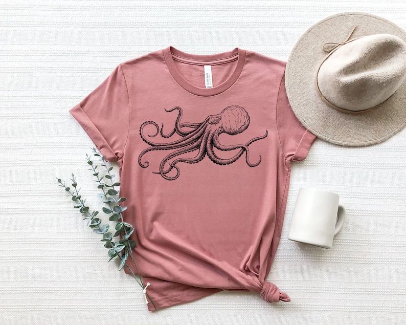 May include: Dusty rose t-shirt featuring a detailed black octopus illustration. The shirt is styled with a beige hat and a white mug. A sprig of greenery adds a decorative touch. The t-shirt is knotted at the bottom.