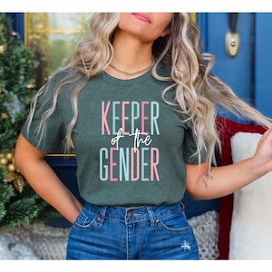 May include: A woman wearing a green t-shirt with the text "KEEPER of the GENDER" in pink and blue lettering. She is wearing blue jeans and has her hair pulled back.