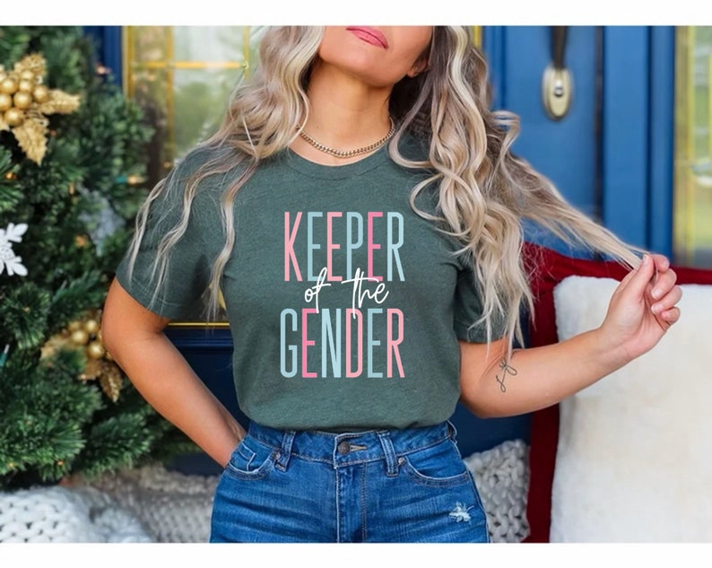 May include: A woman wearing a green t-shirt with the text "KEEPER of the GENDER" in pink and blue lettering. She is wearing blue jeans and has her hair pulled back.