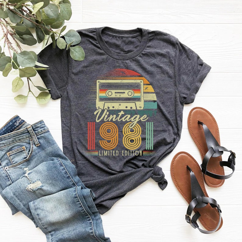 May include: Dark gray t-shirt featuring a retro cassette tape design with the text "Vintage 1981 Limited Edition." The design incorporates orange, yellow, and teal colors. The shirt is paired with blue jeans and black sandals.