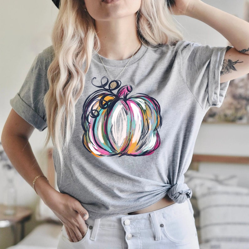 Może przedstawiać: Szary melanżowy t-shirt z kr&oacute;tkim rękawem, ozdobiony centralnym nadrukiem dyni w malarskim stylu, wykonanym wyrazistymi pociągnięciami pędzla w żywych odcieniach magenty, turkusu, pomarańczu, bieli i czerni, z widocznym zakręconym ogonkiem.