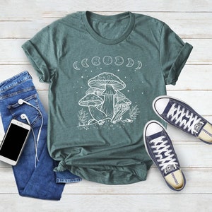 May include: A teal green t-shirt with a white outline of mushrooms and a crescent moon design. The shirt has a crew neck and short sleeves.