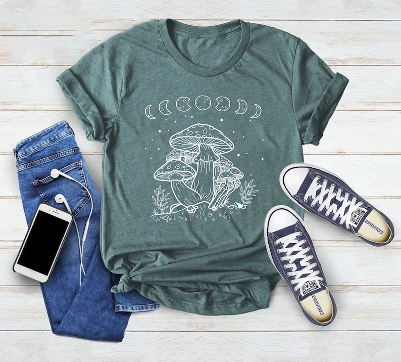 May include: A teal green t-shirt with a white outline of mushrooms and a crescent moon design. The shirt has a crew neck and short sleeves.