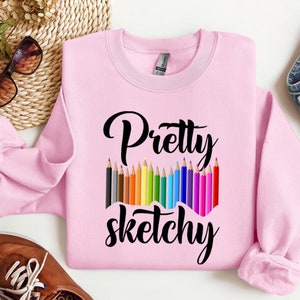以下が含まれることがあります： ピンクのトレーナーに、黒い文字で「Pretty Sketchy」と書いてあります。テキストの下には、カラフルな鉛筆が並んでいます。
