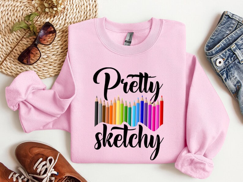 以下が含まれることがあります： ピンクのトレーナーに、黒い文字で「Pretty Sketchy」と書いてあります。テキストの下には、カラフルな鉛筆が並んでいます。