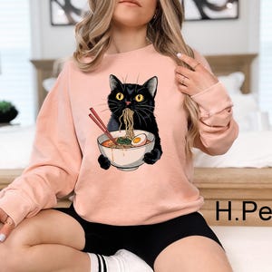 May include: A pink sweatshirt with a black cat eating ramen noodles. The cat is wearing a white bowl with a red egg on top. The text "H.Peach" is visible in the bottom right corner of the image.