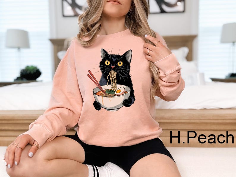 May include: A pink sweatshirt with a black cat eating ramen noodles. The cat is wearing a white bowl with a red egg on top. The text "H.Peach" is visible in the bottom right corner of the image.