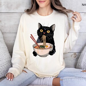 May include: A black cat wearing a white long-sleeved shirt with a graphic of a black cat eating ramen noodles from a bowl. The cat is holding the bowl with its paws. The shirt is on a person sitting on a couch.