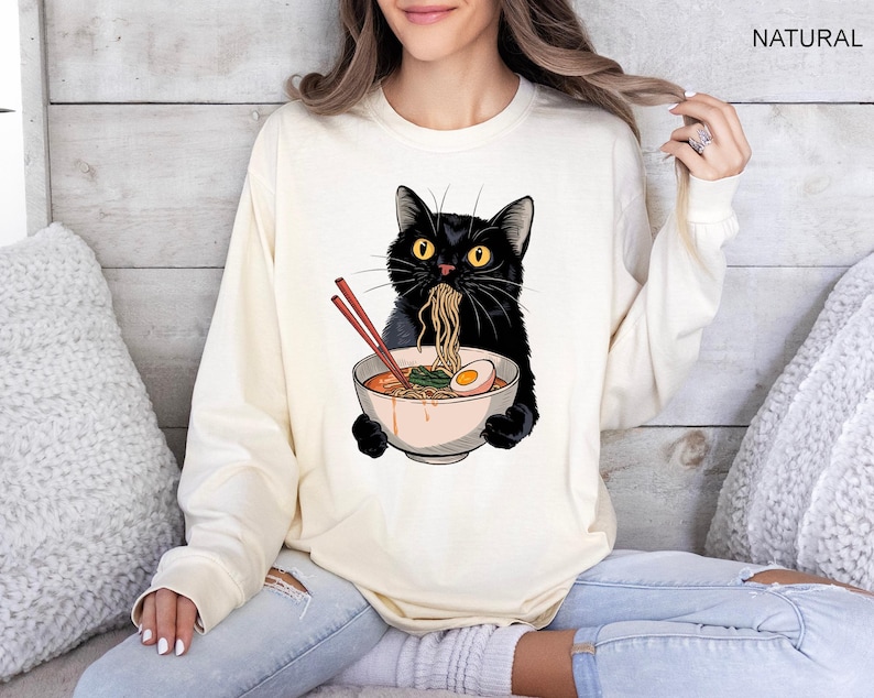 May include: A black cat wearing a white long-sleeved shirt with a graphic of a black cat eating ramen noodles from a bowl. The cat is holding the bowl with its paws. The shirt is on a person sitting on a couch.