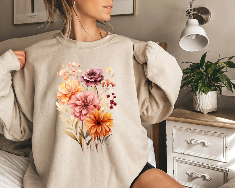 May include: A beige crewneck sweatshirt featuring a central graphic print of a vibrant floral bouquet. The illustration displays various flowers in shades of orange, pink, and deep red, accompanied by delicate foliage and stems in a watercolor style.
