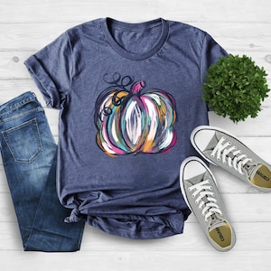 Może przedstawiać: Kr&oacute;tkorękawowy t-shirt w kolorze melanżowego granatu z grafiką abstrakcyjnie namalowanej dyni z widocznymi pociągnięciami pędzla w odcieniach r&oacute;żu, pomarańczu, turkusu i bieli, zestawiony z niebieskimi dżinsami i szarymi niskimi trampkami z pł&oacute;tna.