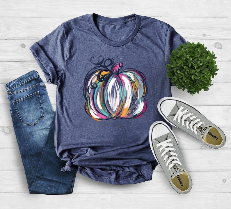 Może przedstawiać: Kr&oacute;tkorękawowy t-shirt w kolorze melanżowego granatu z grafiką abstrakcyjnie namalowanej dyni z widocznymi pociągnięciami pędzla w odcieniach r&oacute;żu, pomarańczu, turkusu i bieli, zestawiony z niebieskimi dżinsami i szarymi niskimi trampkami z pł&oacute;tna.