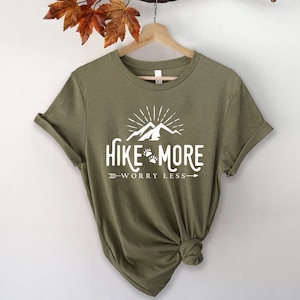 May include: Olive green t-shirt with the white text "Hike More Worry Less" featuring a mountain and paw print design. The shirt has rolled-up sleeves and is hanging on a wooden hanger.