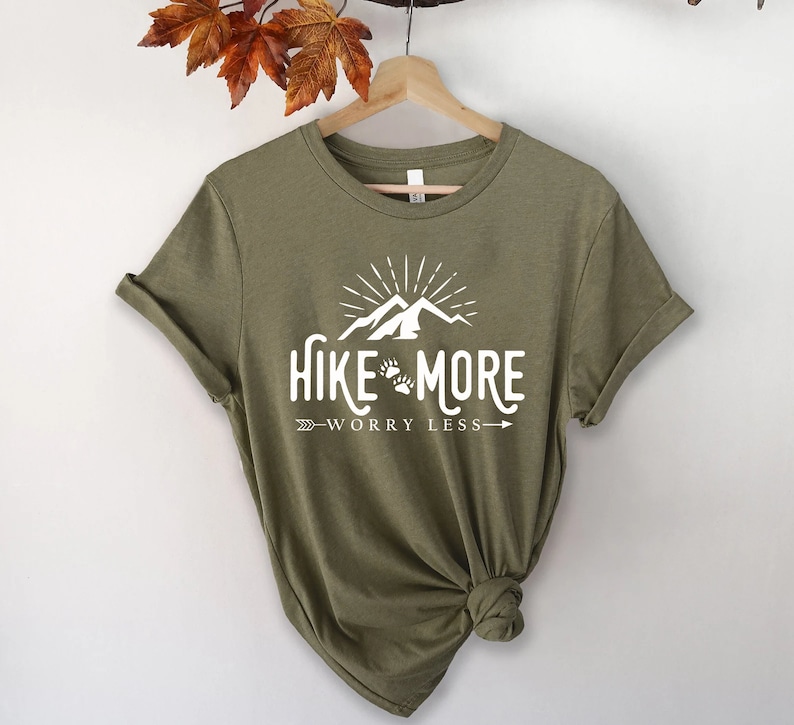 May include: Olive green t-shirt with the white text "Hike More Worry Less" featuring a mountain and paw print design. The shirt has rolled-up sleeves and is hanging on a wooden hanger.