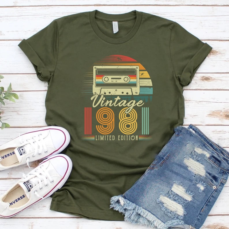 May include: Olive green t-shirt with a retro design featuring a cassette tape, a sunset, and the text "Vintage 1981 Limited Edition." The shirt is paired with white sneakers and denim shorts.
