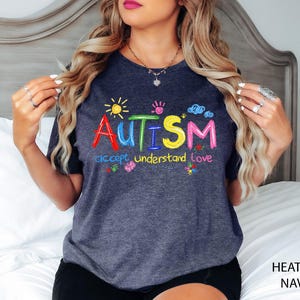 May include: Heather navy t-shirt with the word "AUTISM" in colorful embroidered letters, along with the phrases "accept understand love" and small graphics of a sun, handprints, clouds, and flowers.