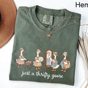 May include: Hemp-colored t-shirt with a graphic of geese dressed in vintage clothing. The geese are depicted shopping and looking in a mirror. The text "just a thrifty goose" is printed below the graphic. A silver necklace is draped across the shirt.