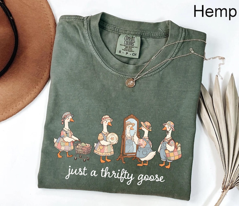 May include: Hemp-colored t-shirt with a graphic of geese dressed in vintage clothing. The geese are depicted shopping and looking in a mirror. The text "just a thrifty goose" is printed below the graphic. A silver necklace is draped across the shirt.