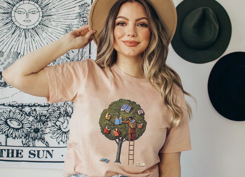 Pu&ograve; includere: Una maglietta color pesca con un'immagine di un albero con libri come foglie. Una persona sta salendo su una scala per raggiungere i libri. Il testo "THE SUN." &egrave; visibile sullo sfondo.
