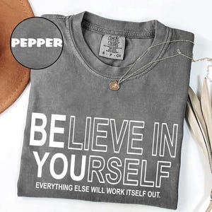 May include: A gray Comfort Colors t-shirt with the word "PEPPER" and the text "BELIEVE IN YOURSELF" in white. The shirt also has the phrase "EVERYTHING ELSE WILL WORK ITSELF OUT." A silver necklace with a pendant is draped on the shirt.