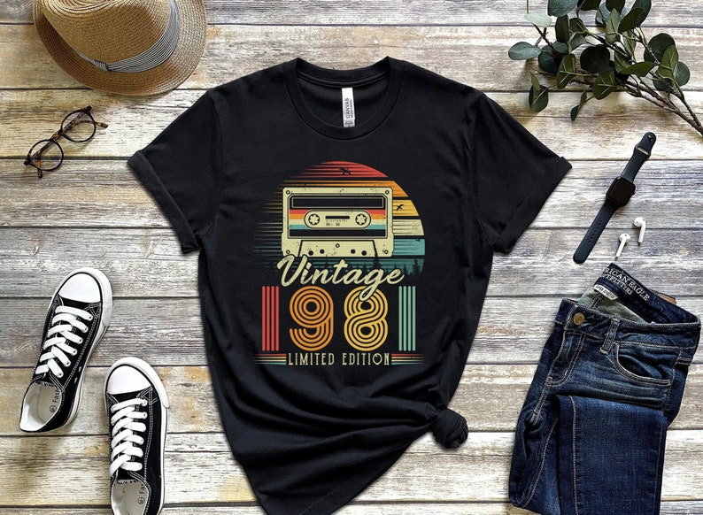 May include: Black t-shirt with a retro design featuring a cassette tape, a sunset, and the text "Vintage 1981 Limited Edition." The design uses orange, yellow, green, and blue colors. The shirt is displayed on a wooden surface with other accessories.