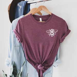 May include: A maroon short-sleeved t-shirt with a white graphic of a bee on the upper left chest. The shirt features a crew neckline and is tied in a knot at the bottom center. The fabric has a heathered texture and is displayed on a wooden hanger.