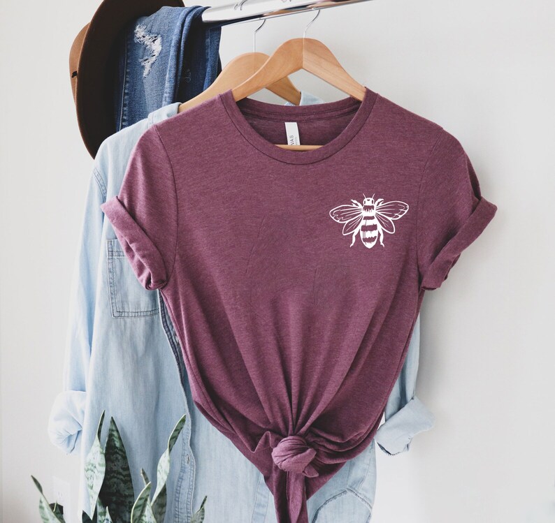 May include: A maroon short-sleeved t-shirt with a white graphic of a bee on the upper left chest. The shirt features a crew neckline and is tied in a knot at the bottom center. The fabric has a heathered texture and is displayed on a wooden hanger.