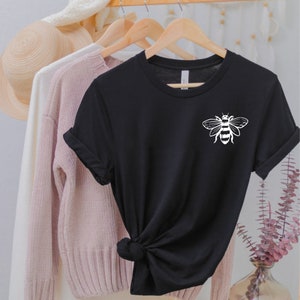 May include: A black short-sleeved t-shirt featuring a white graphic of a bee on the upper left chest. The shirt has a crew neckline and is displayed on a wooden hanger, with the hem tied in a knot at the front to create a gathered appearance.