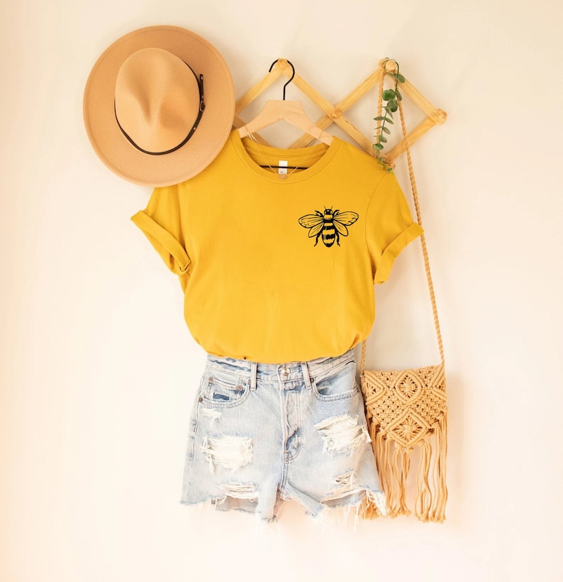 May include: A mustard yellow short-sleeve t-shirt featuring a black line-art bee graphic on the chest, paired with light blue distressed denim shorts, a tan wide-brim hat, and a beige macrame fringe crossbody bag with a wooden wall hanger.