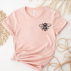 May include: A light pink short-sleeved t-shirt featuring a black graphic of a bee on the upper left chest area. The shirt has a crew neckline and a standard fit, with the hem gathered and tied in a knot at the bottom left side of the garment.