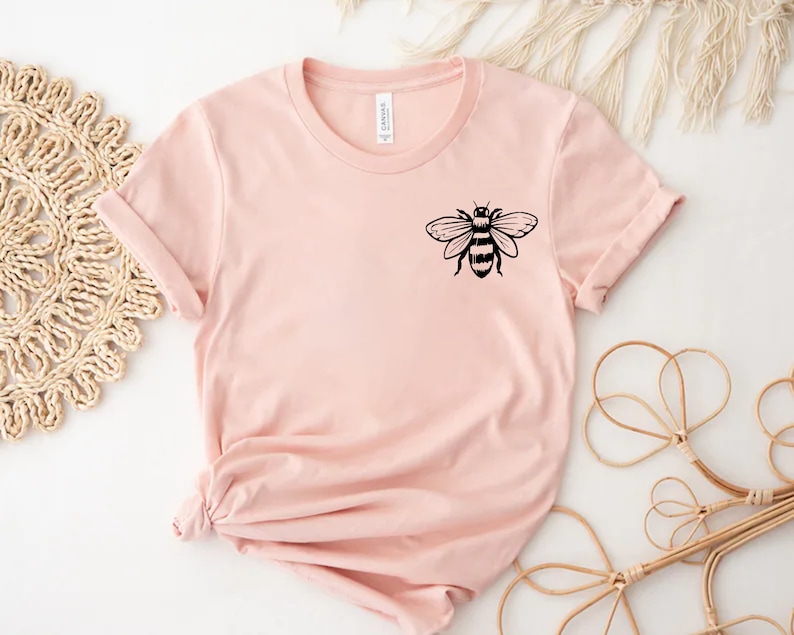 May include: A light pink short-sleeved t-shirt featuring a black graphic of a bee on the upper left chest area. The shirt has a crew neckline and a standard fit, with the hem gathered and tied in a knot at the bottom left side of the garment.