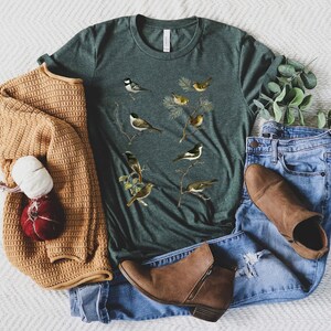 May include: A dark green t-shirt featuring a vintage-style illustration of various birds perched on branches. The shirt is styled with blue jeans, a tan sweater, and brown ankle boots, creating a casual, everyday look.
