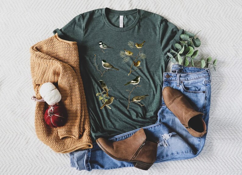 May include: A dark green t-shirt featuring a vintage-style illustration of various birds perched on branches. The shirt is styled with blue jeans, a tan sweater, and brown ankle boots, creating a casual, everyday look.