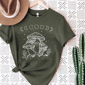 May include: A green t-shirt with a white graphic of mushrooms and a crescent moon design. The shirt has short sleeves and a crew neck.