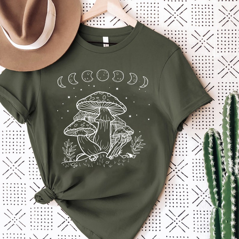 May include: A green t-shirt with a white graphic of mushrooms and a crescent moon design. The shirt has short sleeves and a crew neck.