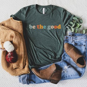 May include: A casual flat lay featuring a dark green t-shirt with the text "be the good" in retro colors, paired with blue jeans, brown ankle boots, and a tan sweater. A decorative pumpkin and sprigs of greenery complete the scene.