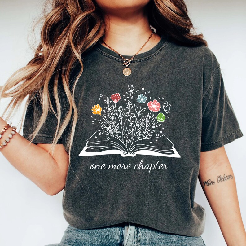 May include: A dark gray t-shirt with a white graphic of an open book with colorful flowers growing out of it. The text "one more chapter" is printed below the book.