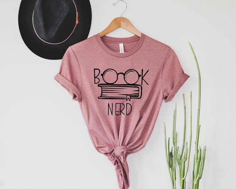 May include: Mauve t-shirt with the words "BOOK NERD" in black, along with a graphic of glasses over a stack of books. The shirt is hanging on a wooden hanger against a white background. A black hat is visible in the background.