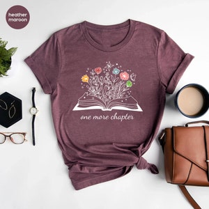 May include: A heather maroon t-shirt with a white graphic of an open book with colorful flowers growing out of it. The text "one more chapter" is printed below the book.