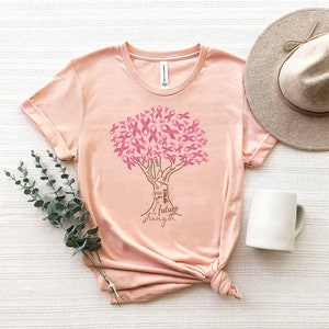 May include: A peach-colored t-shirt featuring a graphic of a tree where the leaves are composed of pink awareness ribbons. The trunk of the tree contains the words cure, hope, faith, grace, future, and strength written in a dark brown script.