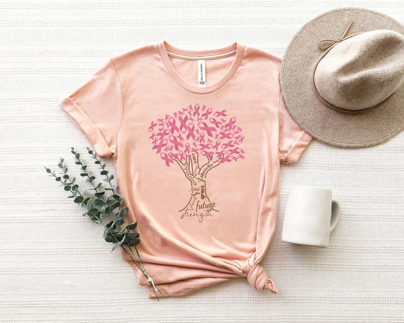May include: A peach-colored t-shirt featuring a graphic of a tree where the leaves are composed of pink awareness ribbons. The trunk of the tree contains the words cure, hope, faith, grace, future, and strength written in a dark brown script.