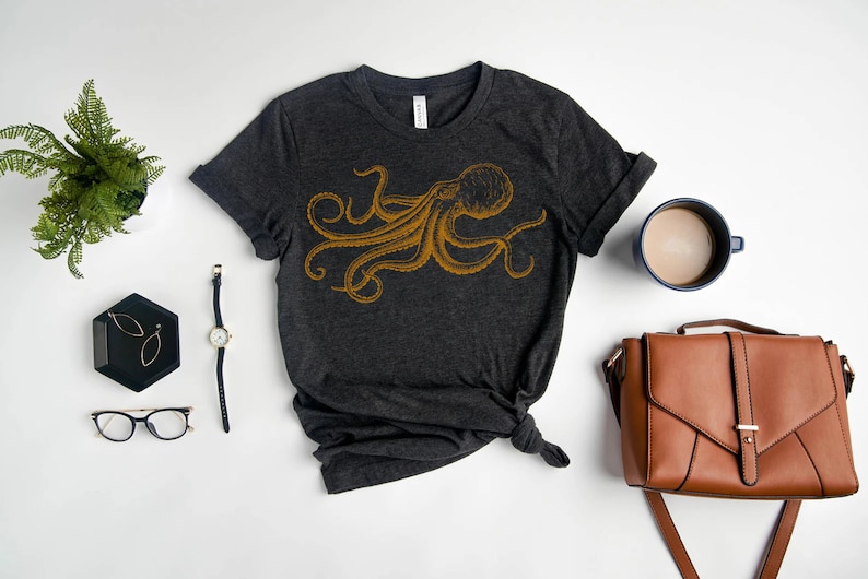 May include: Dark gray t-shirt with a gold octopus design. The shirt is styled with a rolled sleeve and a knot at the bottom. Accessories include a brown purse, a blue mug with coffee, and a black tray with jewelry.
