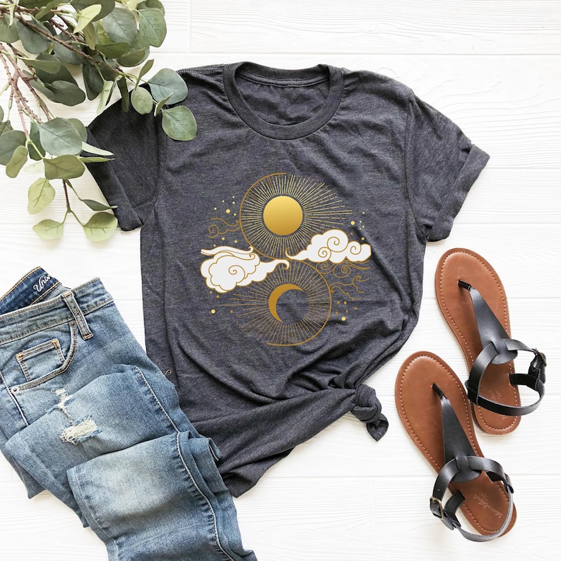 May include: A dark gray t-shirt with a gold sun and moon design. The sun and moon are surrounded by clouds and stars.