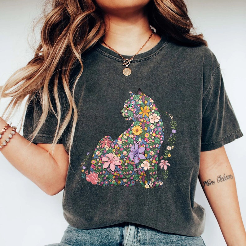 May include: Dark gray t-shirt featuring a floral cat design. The cat is composed of colorful flowers in shades of pink, yellow, purple, and green. The shirt has a vintage, washed look.