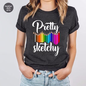 以下が含まれることがあります： ヘザーダークグレーのTシャツに、白い文字で「Pretty sketchy」と書いてあります。テキストの上に、カラフルな鉛筆が並んでいます。