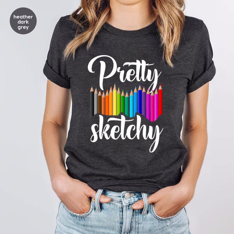 以下が含まれることがあります： ヘザーダークグレーのTシャツに、白い文字で「Pretty sketchy」と書いてあります。テキストの上に、カラフルな鉛筆が並んでいます。